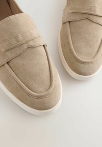 Mocassins à enfiler pour hommes en daim beige avec semelles en caoutchouc blanc sur fond clair.