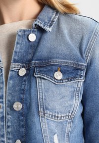 Jeansjacka i ljusblått med blekt tvätt. Har silverknappar, två framfickor och en klassisk krage. Sliten detalj i nederkant.