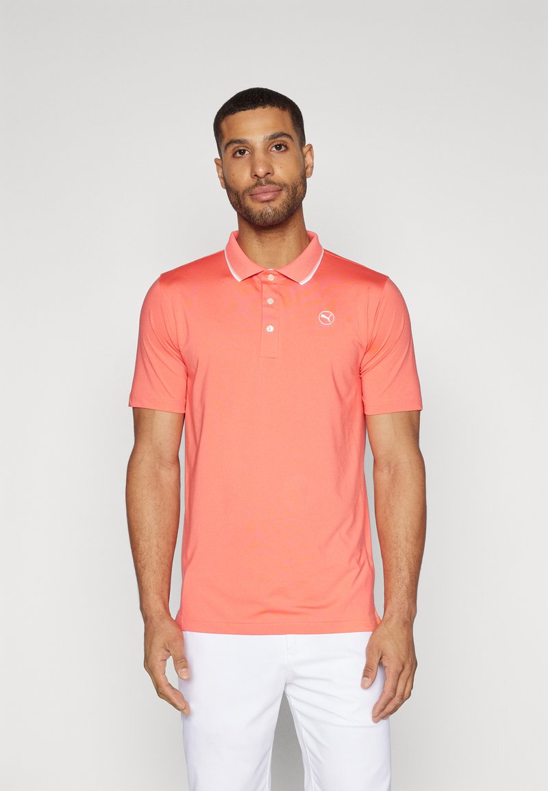 Polo shirt corallo con colletto bianco e logo, maniche corte e design aderente. Indossato con pantaloni bianchi in tessuto liscio.