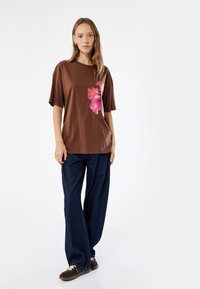 Brun kortærmet T-shirt med et stort pink blomsterprint, parret med løstsiddende mørk denim jeans og brune sko.