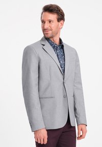 Vaaleanharmaa bleiseri, jossa on kaksipainikeinen etuosa, lovikaulus ja kaksi läppätaskua. Paita on tummassa kukkakuosisessa paidassa. Silkkinen pinta.