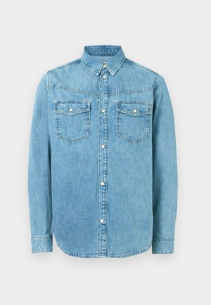 Camisa de denim azul clara com colarinho abotoado, dois bolsos no peito e detalhes de botões prateados. Apresenta mangas longas e um hem em curva.