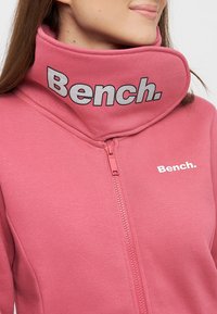 Roze zip-up hoodie met een hoge kraag en een wit mesh-logo. Zachte stof en een textuurdetail bij de kraag verbeteren het ontwerp.