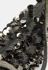 Sort stofpose med en struktur af påsyede skiver og perler. Detaljer inkluderer lukning med lynlås og en kontrasterende sølvkant.