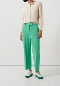 Krämfärgad stickad cardigan med knappslutning framtill; klara gröna skräddarsydda byxor med bälte i midjan; vita sneakers med gröna detaljer.