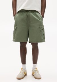 Shorts cargo verts en coton, avec une taille élastique, des poches latérales et une coupe décontractée. Associés à des baskets blanches et des chaussettes.