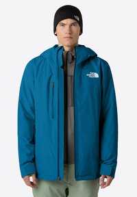 Uomo che indossa una giacca North Face blu sopra una felpa grigia con zip, berretto nero e pantaloni verde chiaro, rivolto verso l'obiettivo su uno sfondo semplice.