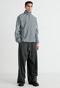 Veste coupe-vent gris clair avec des accents blancs, col montant et poignets élastiques, portée avec un pantalon noir à carreaux ample et des chaussures noires épaisses.