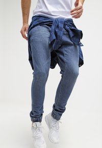 Obepnuté denimové joggery ve střední modři s ribbanými manžetami. Kombinované s uzlem uvázanou tmavomodrou károvanou flanelovou košilí a bílými teniskami.