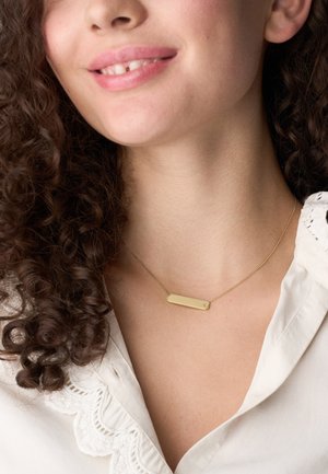 Collier ton or avec un pendentif rectangulaire sur une chaîne délicate. Le pendentif est lisse avec des bords arrondis, porté sur un chemisier blanc.