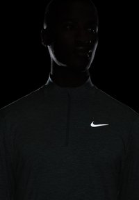 Camisa de manga larga con cremallera de Nike en gris oscuro con un acabado texturizado y logo blanco en la parte superior izquierda, sobre un fondo negro.