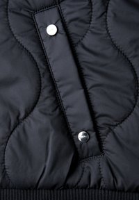 Tissu matelassé noir avec des lignes de couture courbées et deux boutons-pression métalliques brillants sur une bande de fermeture verticale.