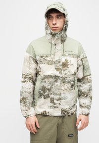 Νεαρός άνδρας που φοράει καφέ-πράσινο καμουφλάζ μπουφάν με κουκούλα και ελιά παντελόνια, κοιτάζοντας προς τα πλάγια απέναντι σε ένα απλό φόντο.