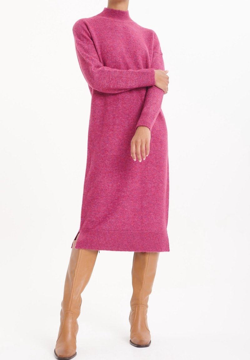 Personne portant une longue robe en tricot rose et des bottes en cuir camel montant jusqu'aux genoux, debout avec un bras croisé sur la poitrine.