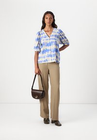 Blå og kremfarget tie-dye-bluse med pufferm, parret med beige bukser og en brun crossover-veske. Teksturen ser glatt ut.