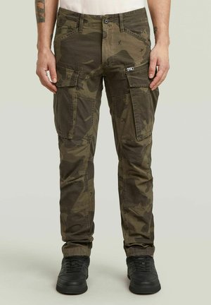 Hombre con pantalones cargo de camuflaje verde con múltiples bolsillos y zapatillas negras, de pie frente a un fondo claro y sencillo.