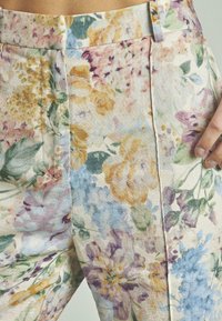 Close-up van bloemige stoffen broek met pastelgele, paarse, blauwe en groene bloempatronen, en een hand die aan de rechterkant rust.