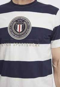 Laivastonsininen ja valkoinen raidallinen t-paita, joka on valmistettu puuvillasta. Siinä on pyöreä kaula-aukko, ympyrälogo laakerinlehtikoristeilla, sekä teksti "1924 SPORTSWEAR".