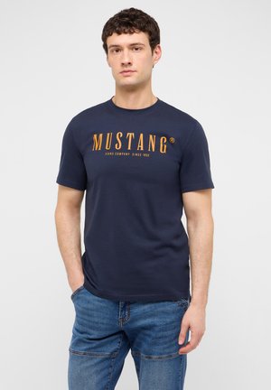 Junger Mann trägt ein dunkelblaues Mustang-T-Shirt mit oranger Schrift und blaue Jeans, steht vor einem schlichten hellen Hintergrund.