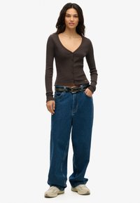 Ung kvinde står med hænderne i lommen, iført en tætsiddende mørkebrun knap-overdel, baggy blå jeans med nagle-bælte og beige sneakers.