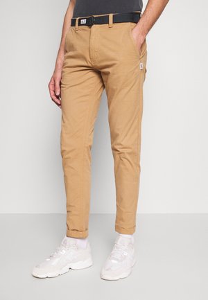 Homme portant un pantalon chino beige avec une ceinture noire, des chaussettes blanches et des baskets blanches, se tenant devant un fond gris clair uni.