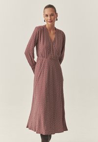 Piros ruha geometriai mintával, hosszú ujjakkal, mély V-nyakkivágással, deréknál megkötve, és lebegő szoknyával; texturált anyag, lágy eséssel.