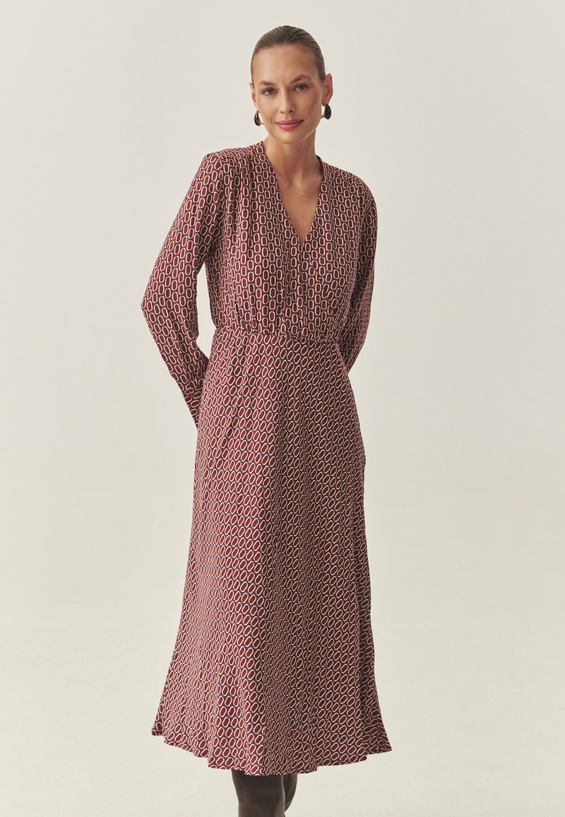 Piros ruha geometriai mintával, hosszú ujjakkal, mély V-nyakkivágással, deréknál megkötve, és lebegő szoknyával; texturált anyag, lágy eséssel.