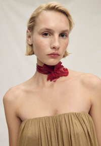 Červená stužková choker s květinovými akcenty nošená kolem krku, v kombinaci s bezramínkovým béžovým topem s záhybovým vzorem pro texturu.