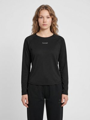 Jeune femme aux cheveux ondulés portant un top de sport noir à manches longues et un pantalon noir, debout devant un fond uni clair.