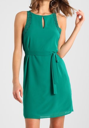 Robe verte avec un décolleté en trou de serrure et des bretelles en dentelle, taille ceinturée, et un tissu léger et lisse. Elle dispose d'une jupe évasée.