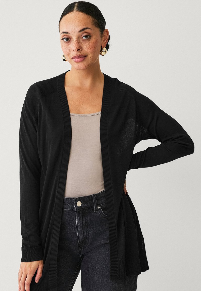 Zwarte open voor cardigan met lange mouwen, ribbelranden, lichte stof, gecombineerd met een lichtbeige top en donkere spijkerbroek.