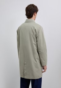 Homme de dos portant un long manteau beige clair avec un col, se tenant devant un fond gris clair uni.