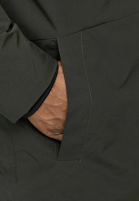 Donkergroene jas van textuurstof, met een zijzak, zichtbare stiksels en een zwarte manchettenvoering. Hand rustend in de zak.