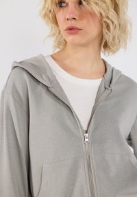 Lys grå zip-up hettegenser laget av myk stoff, med en frontlomme, hette og synlig sølvglidelåsdetalj. Brukt over en hvit skjorte.