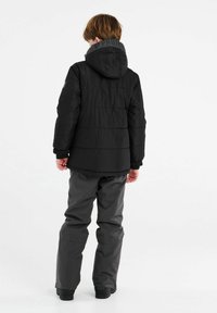 Schwarze Pufferjacke mit Kapuze, gerippten Bündchen und einem Logo am Kragen. Kombiniert mit grauen wasserdichten Hosen und schwarzem Schuhwerk.
