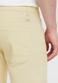 Pantalones de color amarillo claro con una textura suave. Presentan un bolsillo trasero con un detalle de costura y una pequeña etiqueta en la cintura.