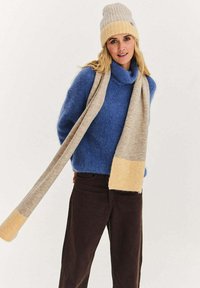 Modrý pletený svetr, béžová šála se žlutými akcenty a béžová čepice. Měkká textura, uvolněný střih a tlumená barevná paleta. Hnědé kalhoty.