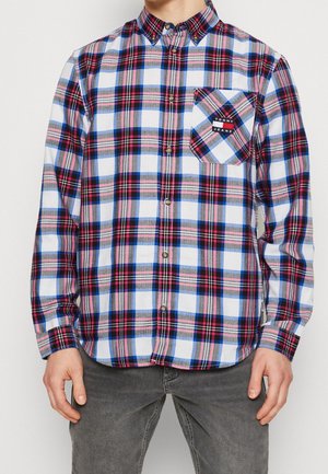 Mann trägt ein blau-weiß-rotes kariertes Hemd mit Knöpfen und einer Brusttasche mit Tommy Jeans-Logo.