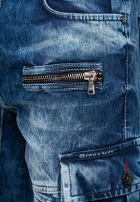Pantaloni cargo in denim con una finitura blu testurizzata, una tasca con zip prominente e un'etichetta di branding. I dettagli strappati arricchiscono il design.