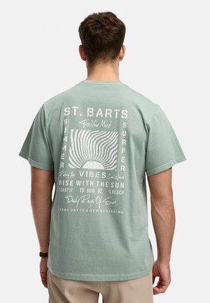 Man wearing light green t-shirt with white sun and wave design and "St. Barts Summer Surfer" text on the back.