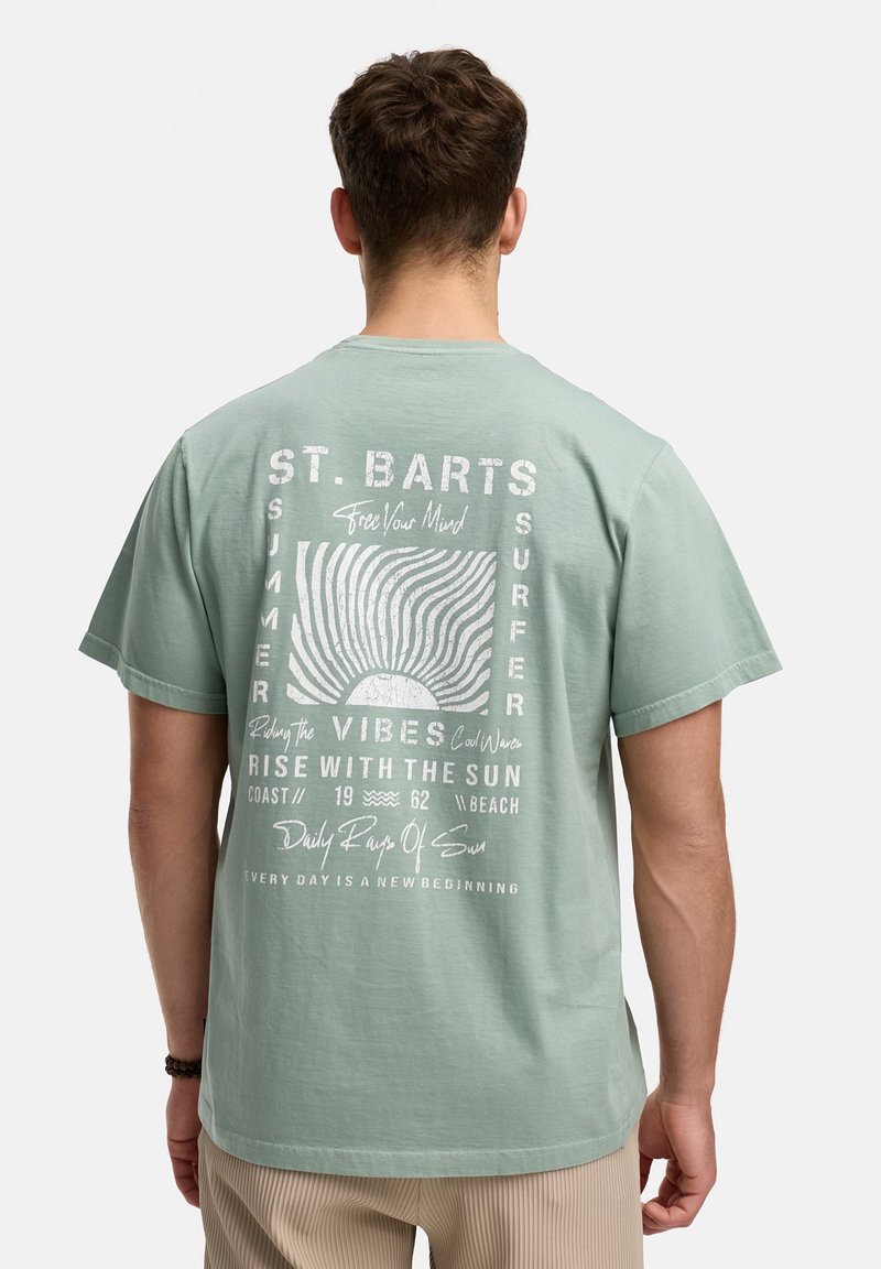 Homme portant un t-shirt vert clair avec un motif blanc de soleil et de vague, ainsi que le texte "St. Barts Summer Surfer" au dos.