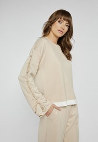 Beżowy sweter z długimi rękawami, z detalami sznurowania wzdłuż rękawów i białą taśmą na dolnym brzegu. Gładka tekstura materiału.