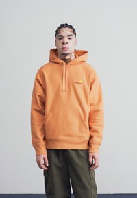 Orange hætte sweatshirt med en kangaroo lomme og logo på brystet, iført med olivengrønne cargo bukser, stående mod en ensfarvet baggrund.