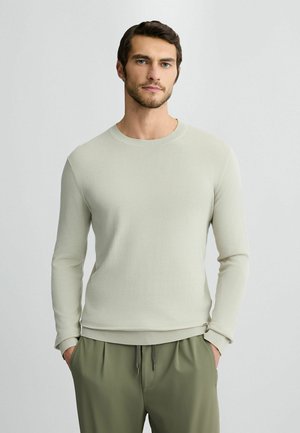 Hellgrauer langärmeliger Pullover mit gerippter Textur und Rundhalsausschnitt, kombiniert mit olivgrünen Hosen, die einen elastischen Bund und einen Kordelzug haben.