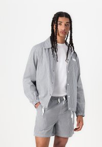 Striped jacket and shorts set in blue and white, featuring a button-up front, elastic cuffs, and drawstring waist. Model wears a white t-shirt.