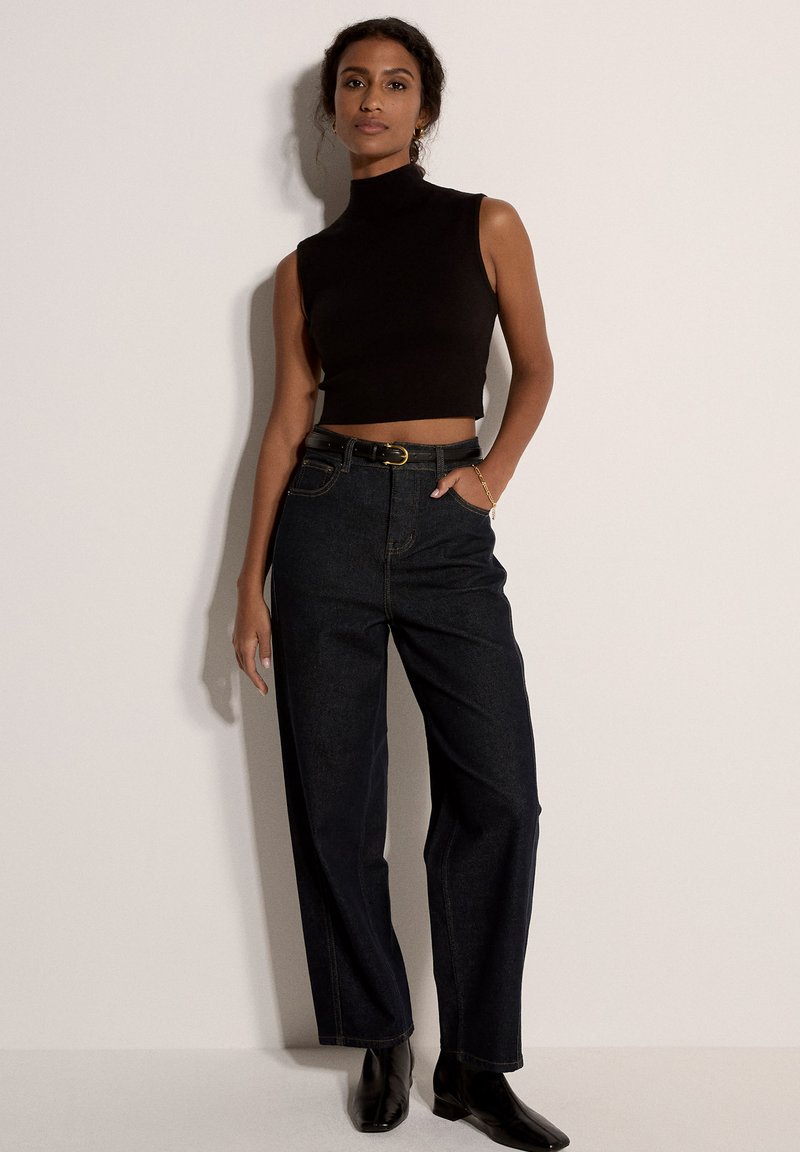 Μαύρο αμάνικο κορμάκι σε στυλ crop top συνδυασμένο με τζιν παντελόνι σε σκούρο μπλε με ψηλή μέση και μαύρη ζώνη. Το μοντέλο φοράει μαύρες μπότες με τακούνι. Απλό φόντο.