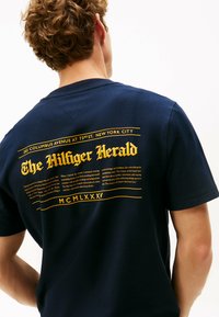 Hombre con cabello rizado que lleva una camiseta azul marino con el texto amarillo "The Hilfiger Herald" y más texto pequeño en la parte de atrás.