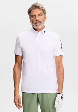 Älterer Mann mit grauem Haar und Bart, der einen weißen Golfhandschuh, ein weißes Poloshirt mit Logo und hellgrüne Hose vor einem schlichten Hintergrund trägt.