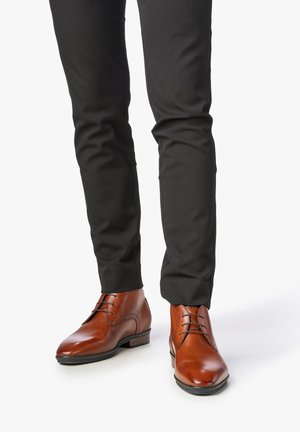 Chaussures de ville en cuir marron au design épuré, avec un bout arrondi et des lacets, associées à un pantalon noir ajusté.