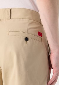 Beige bukser laget av glatt stoff, med en baklomme med en svart knapp og en rød "HUGO"-etikett.